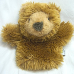 May include: A plush brown teddy bear hand puppet with a light brown face and a dark gray nose. The puppet has a shaggy texture and outstretched arms. A small tag is visible on the side.
