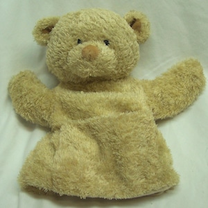 May include: A plush, tan teddy bear hand puppet with a soft, fuzzy texture. The bear has a round head, small ears, a brown nose, and black eyes. The arms are outstretched, and the puppet has a pocket for a hand.