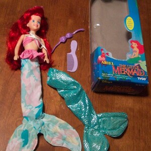 May include: Ariel The Little Mermaid doll with red hair, a pink and blue tail, and a purple comb. The doll is packaged in a blue and white box with the words "Ariel The Little Mermaid" on it.
