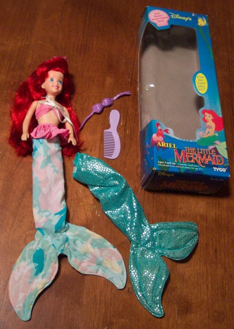 May include: Ariel The Little Mermaid doll with red hair, a pink and blue tail, and a purple comb. The doll is packaged in a blue and white box with the words "Ariel The Little Mermaid" on it.