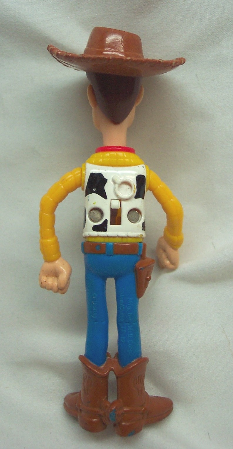 Pu&ograve; includere: Una figurina di plastica di Woody del franchise Toy Story. Woody indossa un cappello da cowboy marrone, una camicia gialla con un bordo rosso, un gilet bianco con un motivo di mucca bianco e nero, jeans blu e stivali da cowboy marroni.