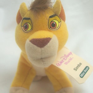 May include: A plush Simba toy from Disney's The Lion King. The toy is yellow with a brown mane, ears, and nose. It has large, expressive eyes and a tag that reads "Walt Disney World" and "Simba Mini Bean".