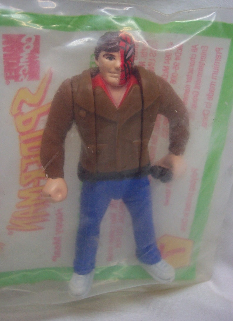 Pu&ograve; includere: Una action figure di plastica di un uomo che indossa una giacca di pelle marrone, jeans blu e una maschera rossa e nera. La figura &egrave; confezionata in un sacchetto di plastica trasparente con uno sfondo verde e giallo. Il sacchetto ha le parole "Spider-Man" e "comics" stampate su di esso.