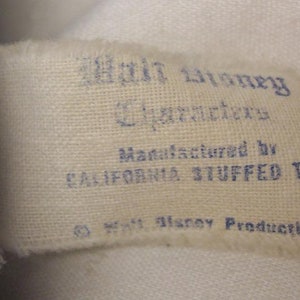 K&ouml;nnte beinhalten: Ein wei&szlig;es Stoff-Etikett mit blauem Text, der "Walt Disney Characters Manufactured by California Stuffed Toys &copy; Walt Disney Productions" lautet.