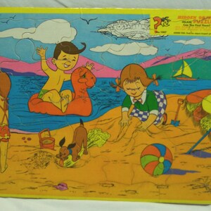May include: A rectangular children's jigsaw puzzle featuring a colorful beach scene with children playing in the sand and water, a small brown dog, a beach ball, and sailboats. The top right corner includes the text Hidden Objects Puzzle.