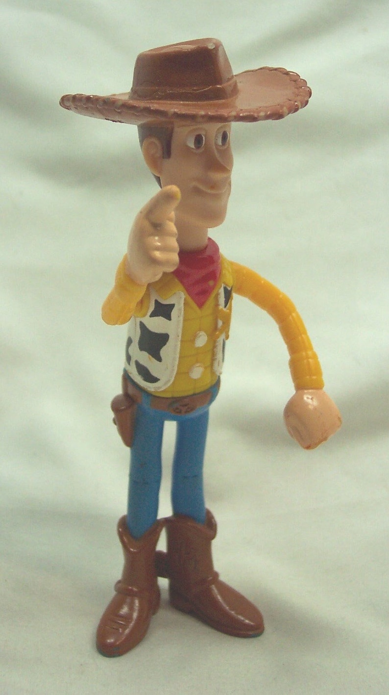 Pu&ograve; includere: Una figurina di plastica di Woody del franchise Toy Story. Woody indossa un cappello da cowboy marrone, un gilet giallo con una bandana rossa e jeans blu. Sta tenendo il braccio destro teso, puntando con l'indice.
