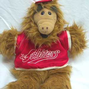 May include: A plush toy of the character ALF, featuring brown fur, a red vest with the word "Arbiters" in white, and a small red hat. The toy has a tan face with black eyes and a brown nose.