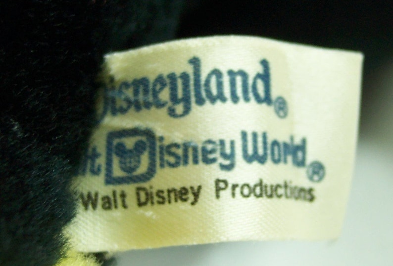 Peut inclure: Une &eacute;tiquette blanche avec du texte bleu qui dit "Disneyland", "Walt Disney World" et "Walt Disney Productions". L'&eacute;tiquette a un petit logo de t&ecirc;te de Mickey Mouse au centre.