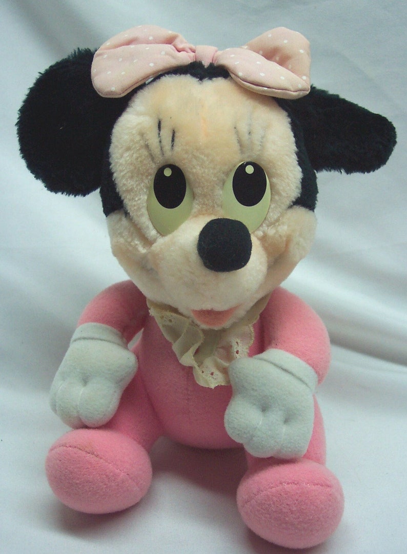 May include: A pink plush toy of Mickey Mouse, wearing a pink and white polka dot bow and a white lace collar. The toy has black ears and a black nose.
