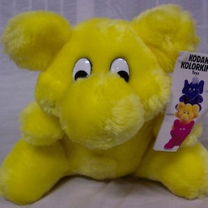 May include: A bright yellow plush toy elephant with large eyes. The elephant is sitting on a white surface. The toy is labeled "Kodak Kolorkins Toys".