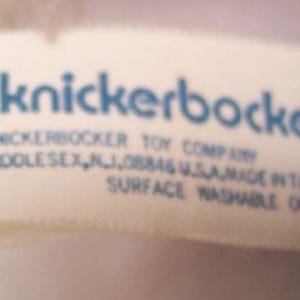 May include: A close-up of a white tag with blue text that reads "knickerbocker". Below the name are the words "KNICKERBOCKER TOY COMPANY MIDDLESEX, NJ, 08846 U.S.A. MADE IN TAIWAN SURFACE WASHABLE ONLY".