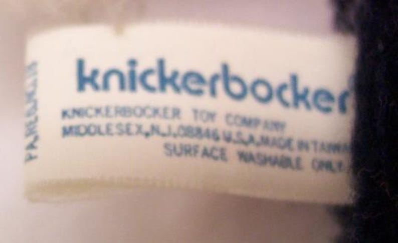 May include: A close-up of a white tag with blue text that reads "knickerbocker". Below the name are the words "KNICKERBOCKER TOY COMPANY MIDDLESEX, NJ, 08846 U.S.A. MADE IN TAIWAN SURFACE WASHABLE ONLY".