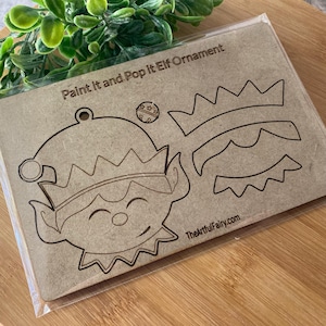 May include: A craft kit for a paint-it-yourself elf ornament. The kit includes a cardboard cutout of an elf face and hat, with additional crown shapes. Text on the packaging reads "Paint it and Pop it Elf Ornament" and "TheArtfulFairy.com".