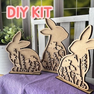 May include: Three wooden bunny cutouts with floral designs, perfect for DIY projects. The bunnies are light brown and stand on small bases. The text "DIY KIT" is displayed in pink letters at the top of the image.