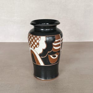 May include: A black ceramic vase featuring a wide, flared rim and a tapered body. The surface is decorated with a hand-painted pattern in white and terracotta brown, consisting of geometric checkerboard squares, curved lines, and abstract shapes.