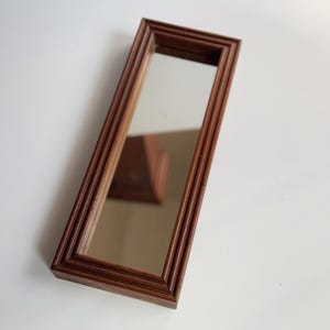 May include: A rectangular, wooden framed mirror with a brown finish. The mirror has a long, narrow shape and a simple, yet elegant design. The frame features a series of parallel grooves, adding texture and visual interest to the piece.