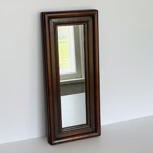 May include: A rectangular, dark brown framed mirror. The frame has a layered design with a beveled edge. The mirror reflects a window with a view of a green lawn and a white window frame. The mirror is leaning against a white wall.