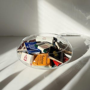 May include: A clear glass bowl filled with a collection of vintage matchboxes. The matchboxes feature various colors and printed text, including "Elk River Town Center Inn," "The Hunt Club," and "Antoine Restaurant," displayed in a shallow, circular container.