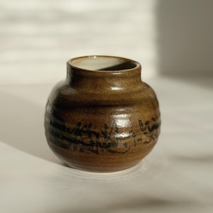 May include: A small, round, brown ceramic vase with a wide opening and a smooth, glossy finish. The exterior features dark, horizontal, hand-painted brushstroke patterns circling the body, contrasting with the earthy, mottled brown glaze.