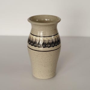 May include: A speckled beige ceramic vase with a flared rim. The vase features a black and white patterned band around the middle. The vase is a decorative item for the home.