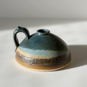 May include: A low-profile ceramic vessel with a single handle and a small central opening. The piece features a multi-colored glaze with bands of deep blue, light blue, tan, and dark brown, with a textured, carved pattern around the lower base.