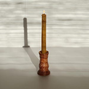 May include: A tall, slender, light brown beeswax taper candle is lit and stands upright in a cylindrical, textured, reddish-brown ceramic candle holder with a woven-like pattern and small white geometric accents around the exterior.