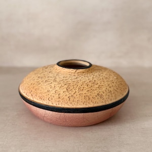 May include: A round, textured ceramic vase with a wide, flat shape. The top portion is a light tan color with a rough, speckled texture. A black band separates the top from the pinkish-red bottom. The opening is black.