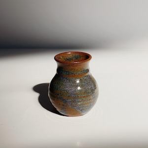 May include: A small, handcrafted ceramic vase with a rounded body and a slightly flared rim. The vase features a speckled, earthy color palette of brown, blue, and orange tones. The interior and rim are a darker brown.