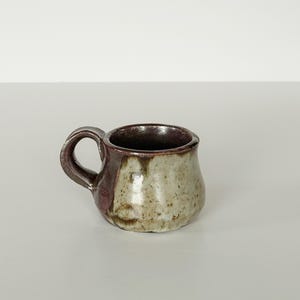 May include: A small, handmade ceramic mug with a rounded body and a handle. The mug features a speckled, off-white exterior with a dark burgundy accent on the handle and side. The interior is a deep brown color.