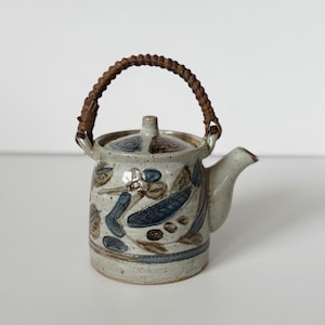 May include: A ceramic teapot with a woven handle. The teapot is off-white with blue and brown floral designs. The spout and lid are light beige. The handle is made of woven brown material.