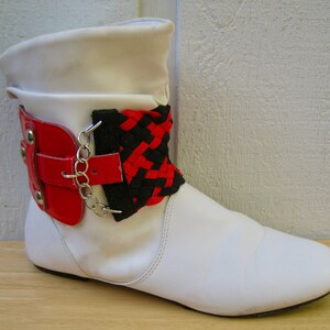 May include: White ankle boots with a red and black braided strap around the ankle. The strap has a silver buckle and chain.