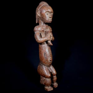 Peut inclure: Une figurine en bois sculpté d'une personne, debout, les bras croisés. La figurine est de couleur marron moyen, avec un grain de bois visible et un aspect vieilli. La figurine a un visage détaillé et est probablement une œuvre d'art tribal africain.