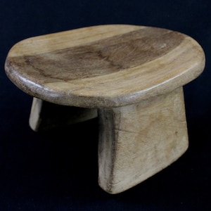 Puede incluir: Un pequeño taburete de madera hecho a mano con un asiento ovalado de color marrón claro. El asiento está sostenido por tres patas rectangulares. La veta de la madera es visible, lo que le da un aspecto rústico. El taburete probablemente se utiliza para sentarse o como objeto decorativo.