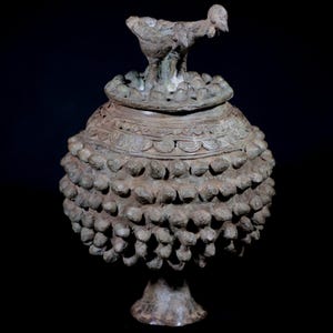 Puede incluir: Un jarrón de bronce ornamentado con tapa, con una escultura de ave en la parte superior. El cuerpo del jarrón presenta un patrón texturizado y redondeado, y se apoya sobre una base de pedestal. El diseño sugiere un objeto antiguo o decorativo.