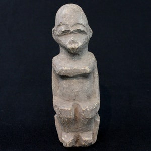 May include: A small, gray stone sculpture of a seated figure with a simplified, stylized form. The figure has a rounded head with a flat face and no visible features.