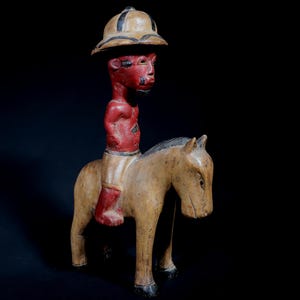 Peut inclure: Sculpture en bois sculpté d'une figurine à cheval. La figurine est peinte en rouge et porte un chapeau beige à rayures noires. Le cheval est beige avec des sabots noirs et une crinière noire.