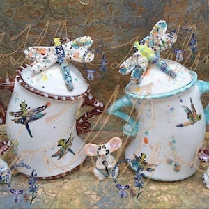 Puede incluir: Dos jarras de cerámica blanca con tapas de libélula y asas marrones y azules. Las jarras están decoradas con pintura salpicada de colores y diseños de libélulas. Cuatro pequeños ratones de cerámica blanca están sentados frente a las jarras.