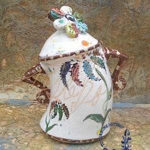 以下が含まれることがあります： 茶色の取っ手とカラフルな花がついた白い陶器の壺。壺は緑、青、茶色の葉と花が描かれた絵柄で飾られています。