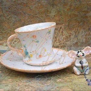 Puede incluir: Una taza de té y un platillo de cerámica blanca con un diseño moteado y una pequeña figurita de ratón de cerámica blanca. La taza de té tiene un asa y un cuenco ancho y poco profundo. El platillo tiene un borde festoneado. La figurita de ratón tiene una cola larga y sostiene un objeto pequeño en sus patas.