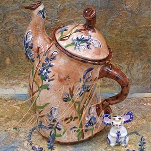 May include: A ceramic teapot with a floral design featuring blue flowers and green leaves. The teapot has a brown handle and a lid with a floral design. A small ceramic mouse figurine is sitting next to the teapot.