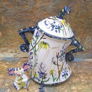 以下が含まれることがあります： 青と黄色の花柄が描かれた白い陶器の壺。青い蓋にはつまみがあり、壺には持ち手が2つついている。壺はざらざらした表面の上に置かれている。壺の前に、黄色の花柄が描かれた小さな白い陶器のネズミが置かれている。