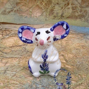 May include: A white ceramic mouse figurine with blue and pink painted details. The mouse is holding a bouquet of blue flowers in its paws.
