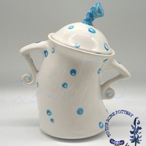 May include: A white ceramic jar with a lid, decorated with blue polka dots and a blue handle. The jar features two curved handles and a whimsical design. The jar has the text "To the Bone Pottery" on the side.