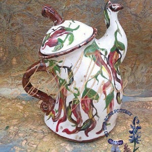 Könnte beinhalten: Eine weiße Teekanne aus Keramik mit einem braunen Henkel und Ausguss. Die Teekanne ist mit einem Blumendesign aus roten, grünen und gelben Blumen verziert. Die Teekanne steht auf einer braunen und weißen strukturierten Oberfläche.