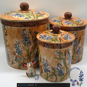 May include: A set of three ceramic storage jars with lids. The jars are tan with brown rims and lids. They feature blue floral designs and green leaves. Each jar has a brown knob on the lid.