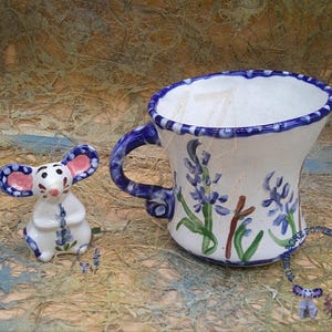 May include: A white ceramic mug with a blue rim and a floral design of blue flowers and green leaves. A small white ceramic mouse figurine with pink ears and blue flowers painted on it is in the foreground.