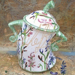 Puede incluir: Una tetera de cerámica blanca con tapa y asa verdes. La tetera está decorada con un patrón floral en tonos rosa, azul y verde.