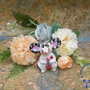May include: A white ceramic mouse figurine with black and white painted details, holding a bouquet of purple flowers. The mouse is surrounded by artificial flowers in shades of peach, white, and blue.