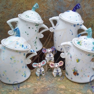 Puede incluir: Cuatro teteras de cerámica blanca con tapas y diseños de pintura salpicada azul y verde. Las teteras están dispuestas en semicírculo con tres pequeñas figuritas de ratones de cerámica blanca delante de ellas.
