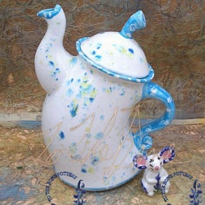 May include: A white ceramic teapot with blue and yellow floral accents and a matching lid. The teapot has a curved handle and spout, and is accompanied by a small mouse figurine. The teapot has the word "Shelly" written on it.